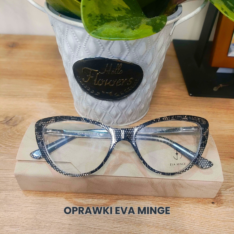 Zdjęcie przedstawia stylowe damskie okulary korekcyjne marki Eva Minge. Oprawki mają kształt typu "kocie oczy" i charakteryzują się czarnym, drobno nakrapianym wzorem na przezroczystym tle. Okulary leżą na beżowym, eleganckim etui, które umieszczone jest na drewnianym blacie. W tle widoczna jest biała doniczka w stylu vintage z zieloną rośliną oraz napis "OPRAWKI EVA MINGE" u dołu grafiki.