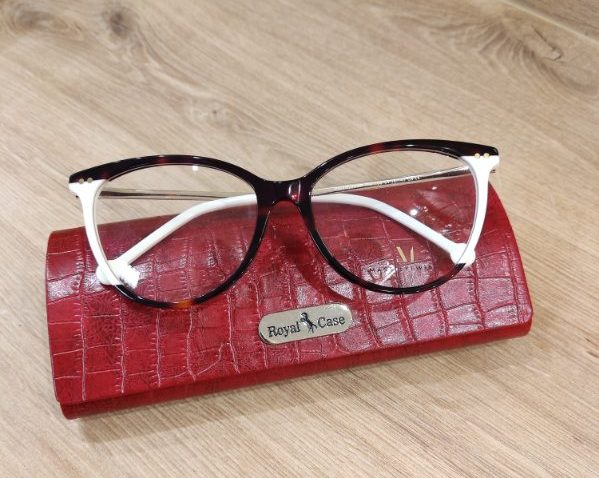 Okulary korekcyjne w oprawkach cat-eye w kolorze szylkretowym z białymi zausznikami i złotymi zdobieniami przy zawiasach, oparte na czerwonym etui na okulary marki Royal Case z fakturą imitującą skórę krokodyla. Tło drewniane w jasnym odcieniu.