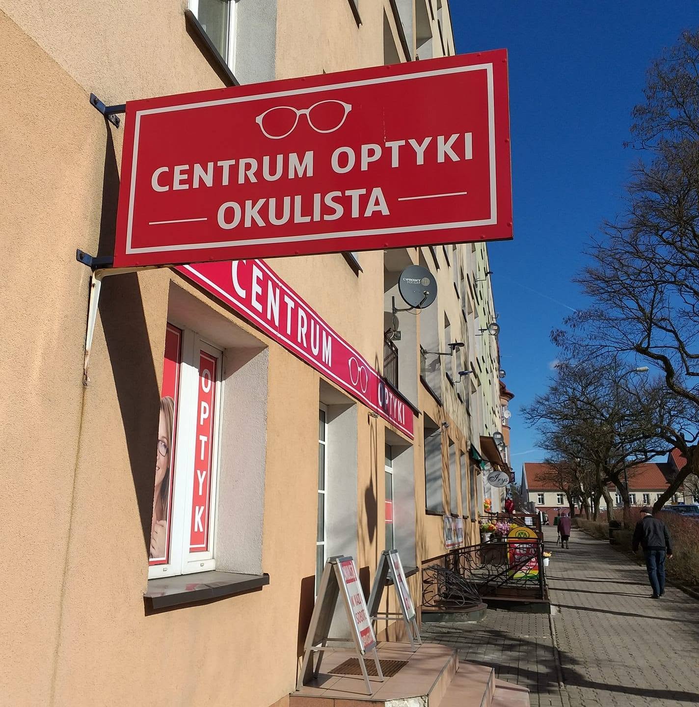 Optyk Olecko Czerwony szyld reklamowy z napisem "CENTRUM OPTYKI OKULISTA" i grafiką okularów, zamontowany na ścianie budynku. W tle widoczny chodnik miejski, idący ludzie oraz bezchmurne, niebieskie niebo.