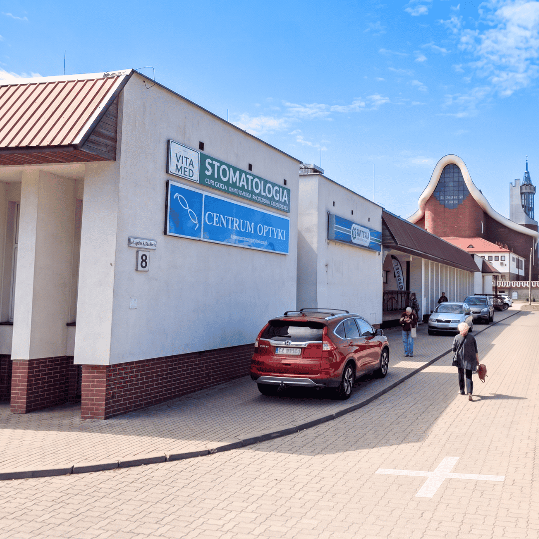 Widok na parterowy budynek usługowy z szyldami gabinetu stomatologicznego „Vita Med” oraz „Centrum Optyki”. Przed budynkiem, na brukowanym chodniku, zaparkowany jest czerwony SUV, a w tle widać nowoczesną bryłę kościoła z ceglaną fasadą pod bezchmurnym, niebieskim niebem Optyk Białystok.
